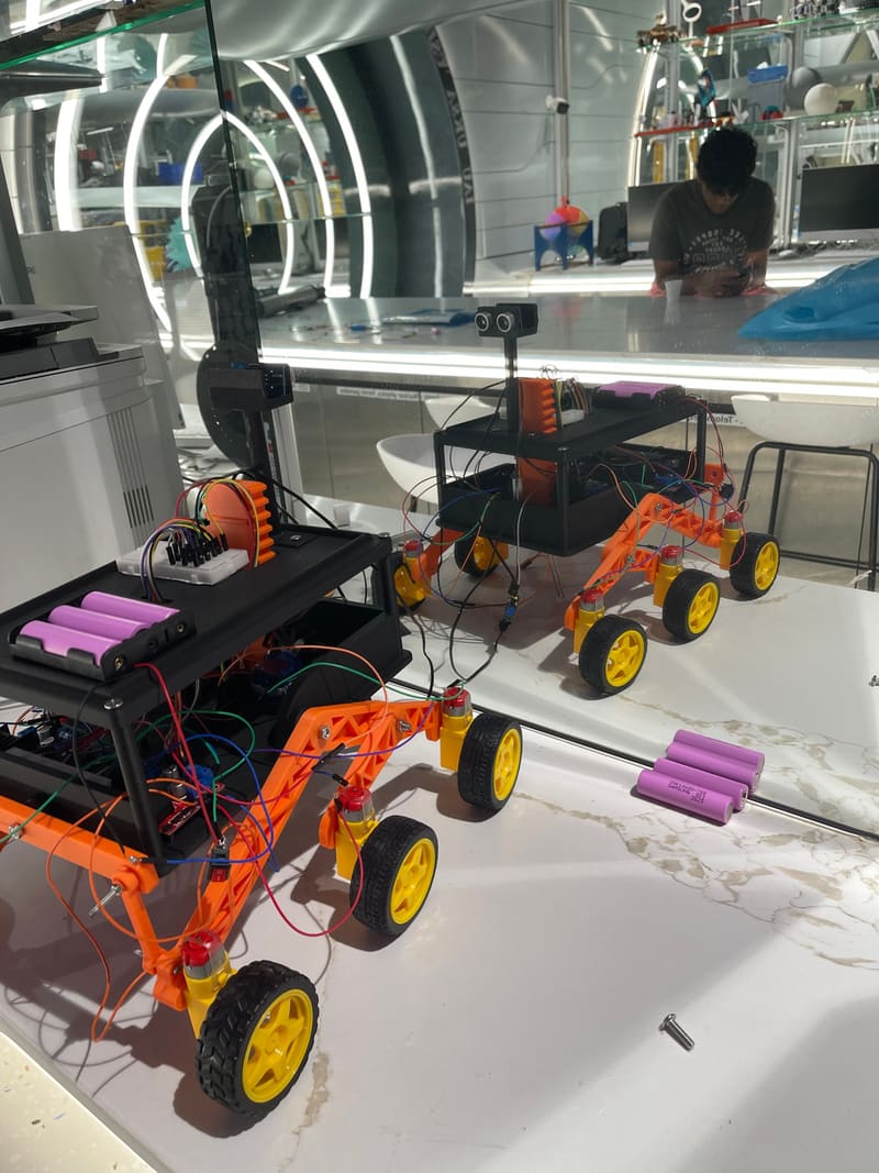 Two Parakh chassis on the makerspace bench mid-assembly, jumper wires everywhere and spare 18650 cells on the table.