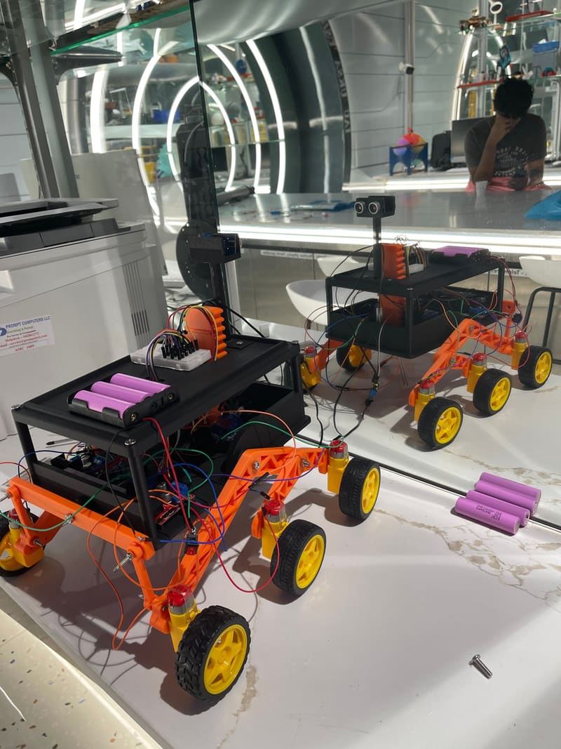 Both rover prototypes side by side on the bench, orange rocker-bogie arms and yellow wheels fitted, batteries waiting.