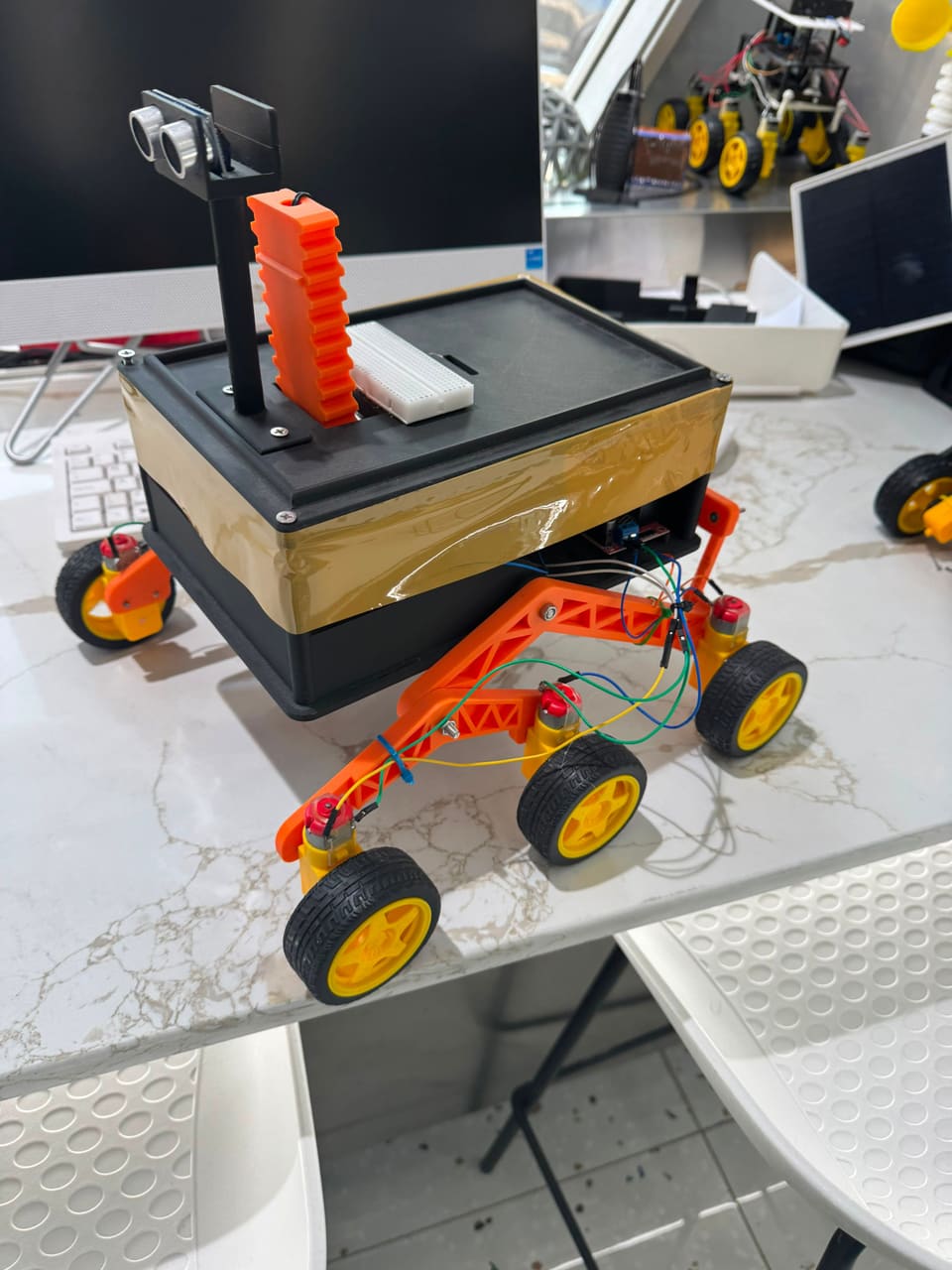 The Parakh rover prototype on a workbench: black 3D-printed chassis wrapped in gold mylar foil, orange rocker-bogie arms, yellow wheels, and a mast carrying twin ultrasonic sensors.