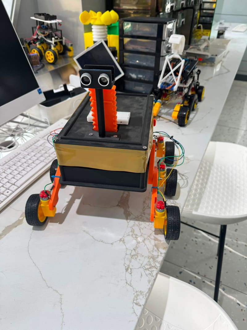 Head-on view of the Parakh rover: twin ultrasonic sensors like a pair of eyes above the gold mylar-wrapped chassis, with a solar panel behind.